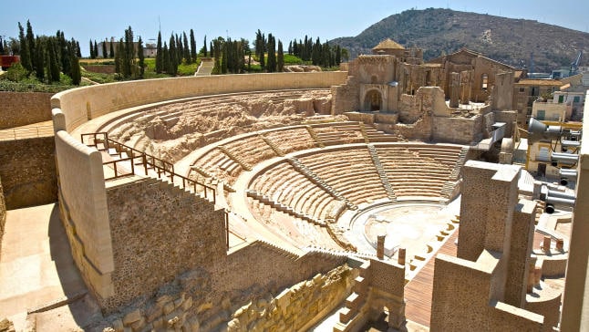 An ancient amphitheater is depicted, showcasing tiered stone seating arranged in a semi-circular layout. The structure features stone arches and walls, with a backdrop of green trees and a distant hillside under a clear blue sky.