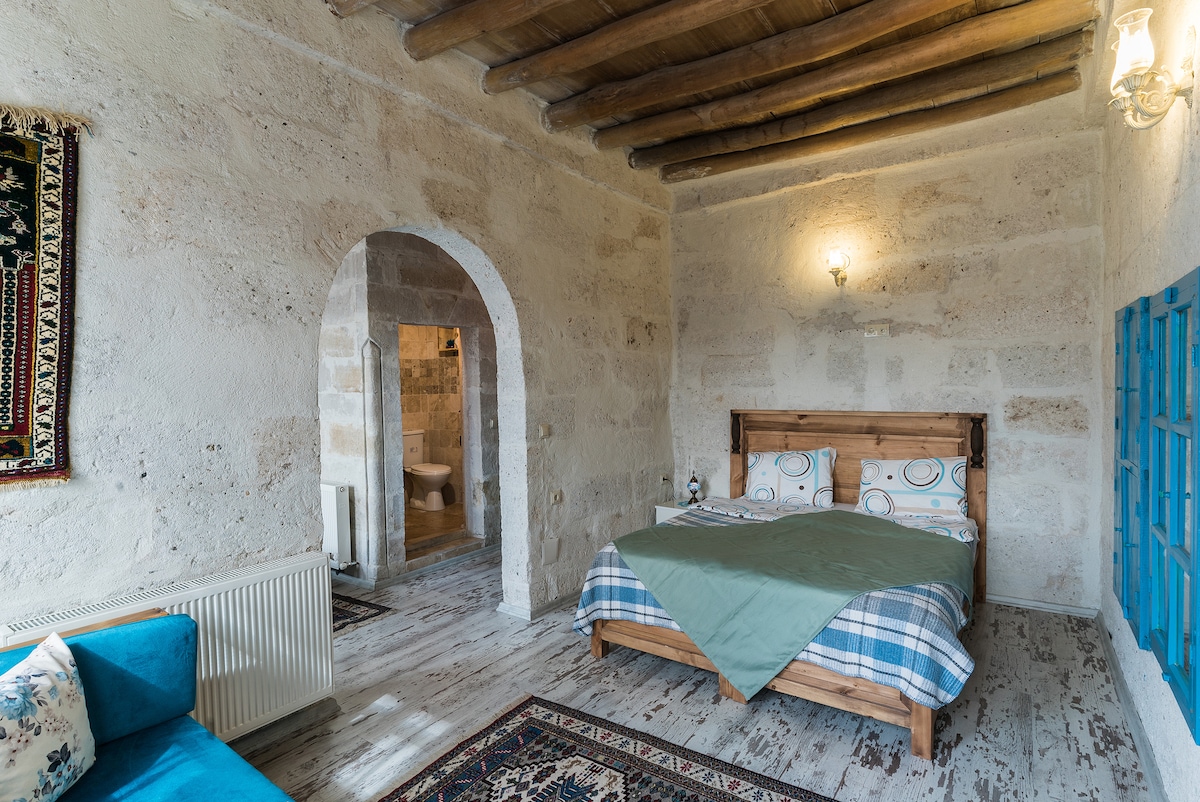 A room features stone walls and wooden beams, creating a rustic ambiance. A spacious bed with patterned bedding is positioned against one wall. A seating area is offered by a blue sofa, and an arched doorway leads to a bathroom in the background.