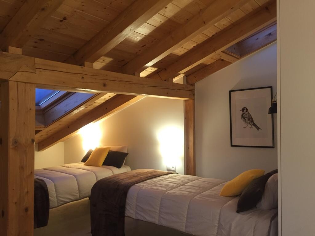 A cozy sleeping area showcases two side-by-side beds, each adorned with soft blankets and decorative pillows. The wooden beams of the ceiling create a warm atmosphere, complemented by soft lighting from wall sconces. A piece of artwork is displayed on the wall, enhancing the serene environment.