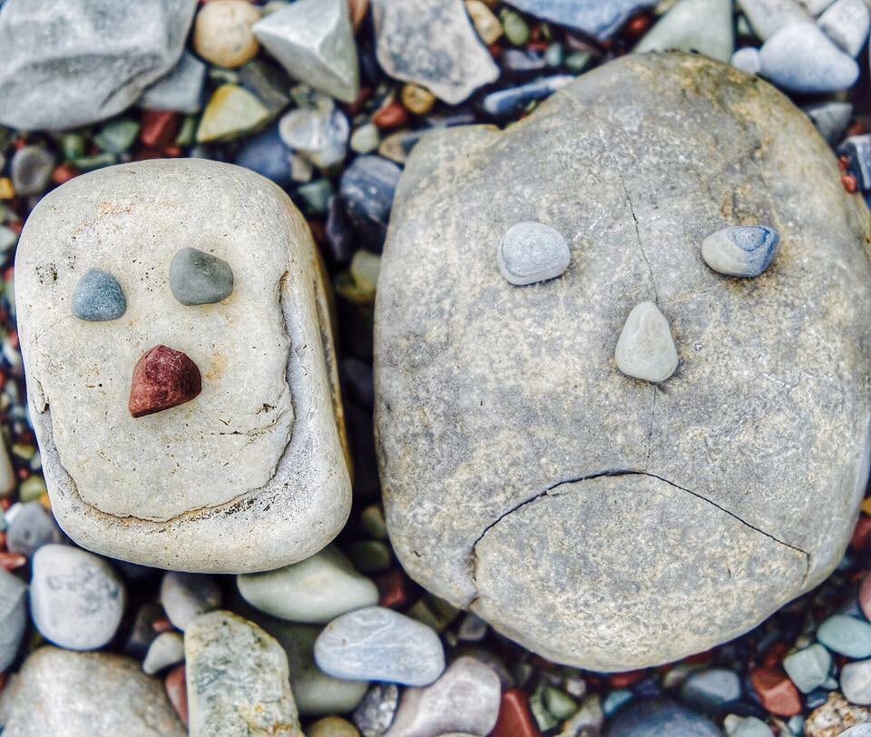 Two rocks are arranged on a bed of smaller pebbles, resembling playful faces. The larger rock features a simple expression with smaller stones for eyes and a frown, while the smaller rock has a smiling face. The surrounding pebbles provide a textured, colorful backdrop.