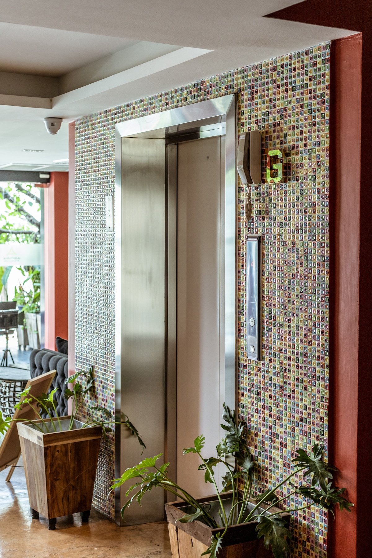An elevator positioned within a vibrant mosaic-tiled wall is displayed. The entrance is accompanied by decorative potted plants, and the surrounding area features a modern atmosphere. Soft lighting enhances the colorful tiles, creating a welcoming environment for guests.