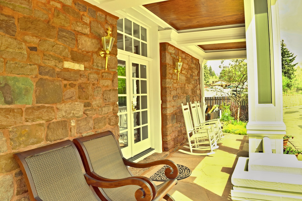 A welcoming front porch features stone walls and sleek wood ceiling accents. Two rocking chairs are positioned for leisure. A pair of French doors opens into the apartment, inviting natural light into the space. Surrounding greenery adds a tranquil element.