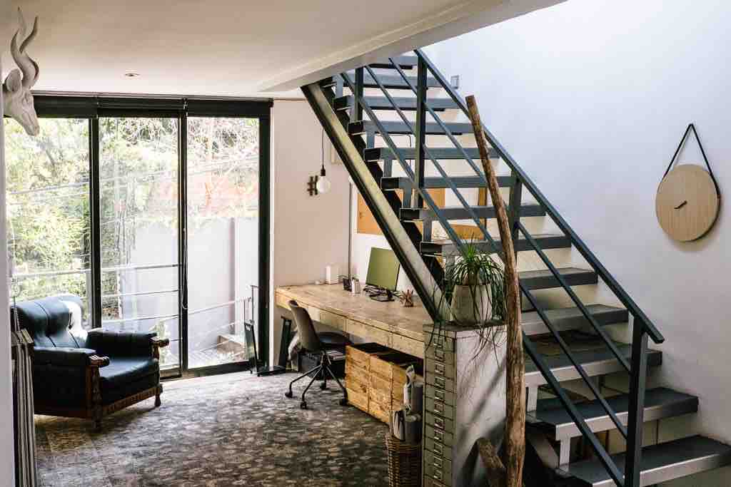 A bright and airy workspace is featured on the middle floor, with a wooden desk, green office chair, and rustic storage units. A comfortable armchair is positioned nearby, and large glass doors provide natural light and a view of the outdoor area. A staircase leads to the upper floors.