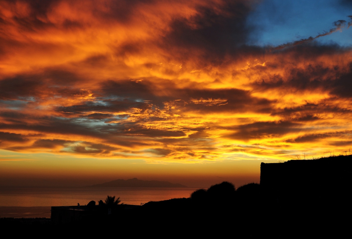 A stunning sunset is captured with vibrant hues of orange, yellow, and blue blending in the sky. Silhouettes of distant hills and coastal outlines gently frame the scenic view, providing a serene backdrop to the tranquil waters below.