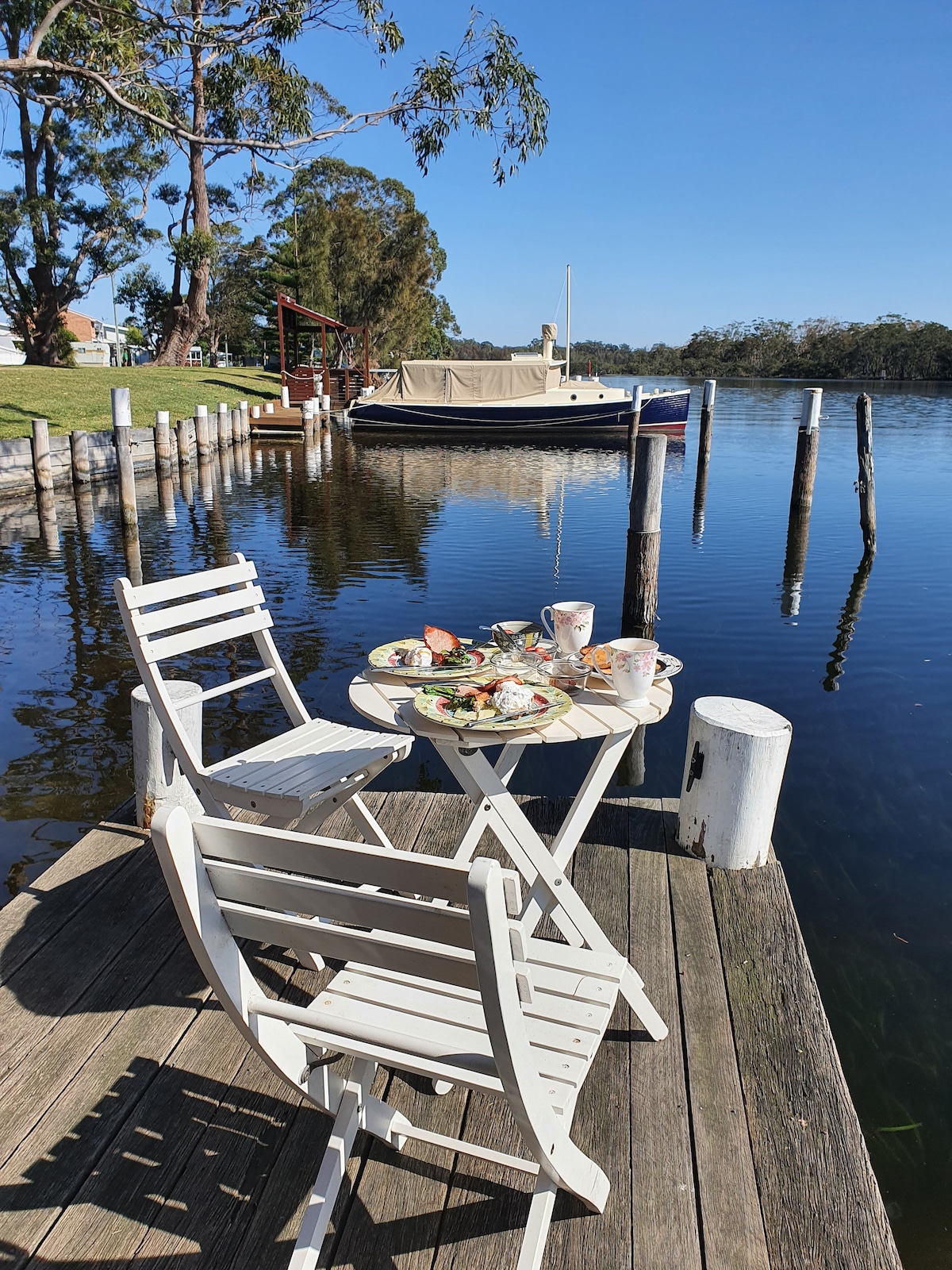 A small white table is set with plates of food and cups, positioned near the water's edge. Two white folding chairs are visible. A boat is moored nearby, while tall trees line the opposite shore, creating a serene waterfront scene.