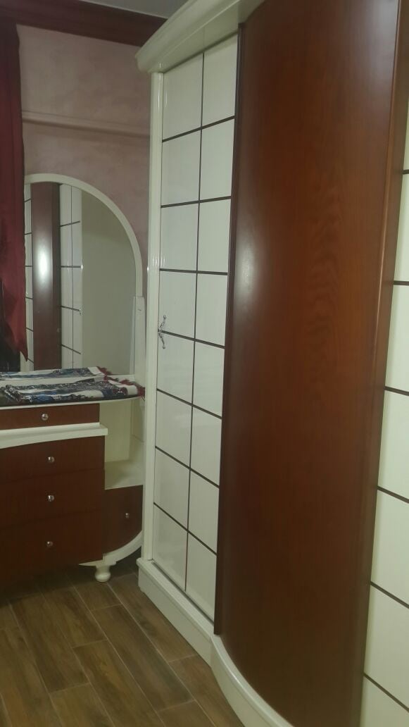 A wooden wardrobe with a sleek design stands alongside a matching dresser. The wardrobe features a mirrored panel and a minimalist style, while the dresser has several small drawers for storage. Soft lighting enhances the warm tones of the wood.