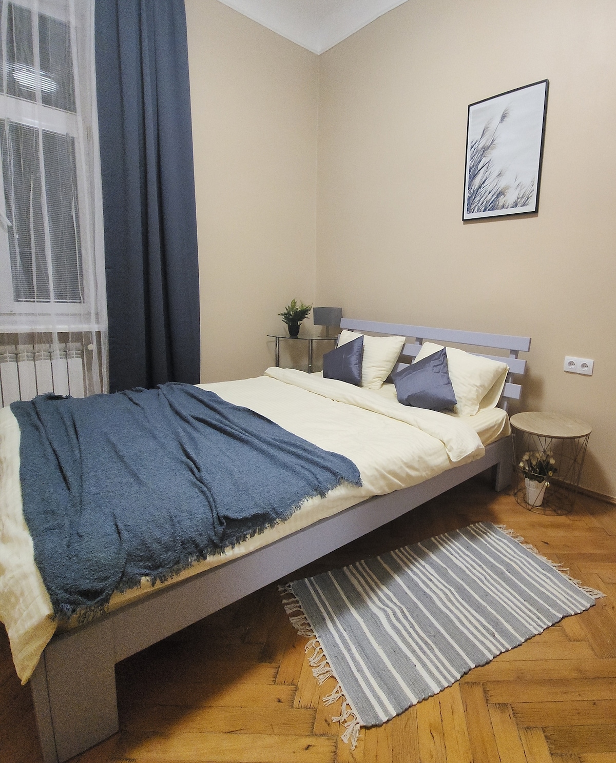 A calming bedroom features a spacious bed dressed with soft linens and decorative pillows. A light blue throw rests atop the bed, while a woven rug adds texture to the wooden floor. Natural light filters through a window dressed with dark curtains.