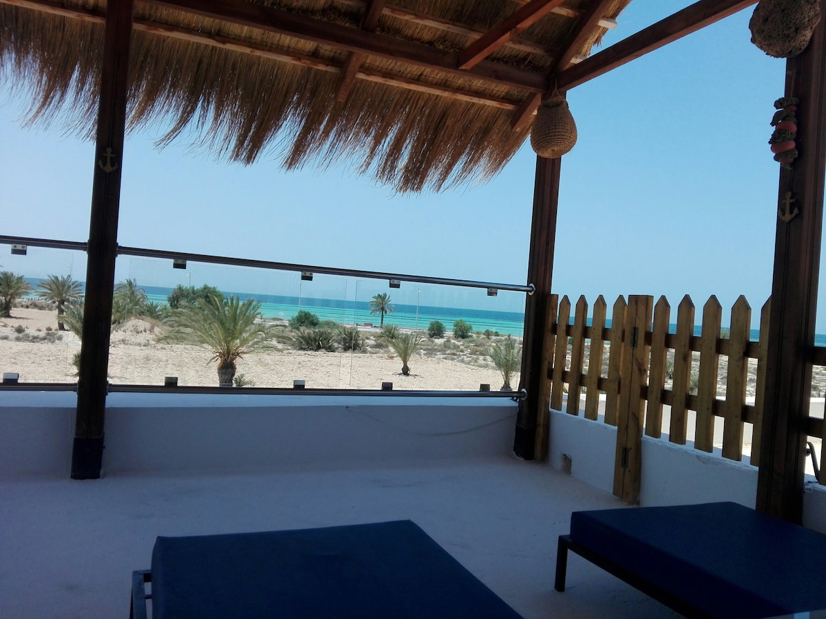 An outdoor terrace features a thatched roof and provides views of the sandy landscape and ocean. Two loungers are positioned for relaxation, while a wooden fence enhances privacy. Palm trees are visible in the distance, contributing to the serene coastal ambiance.