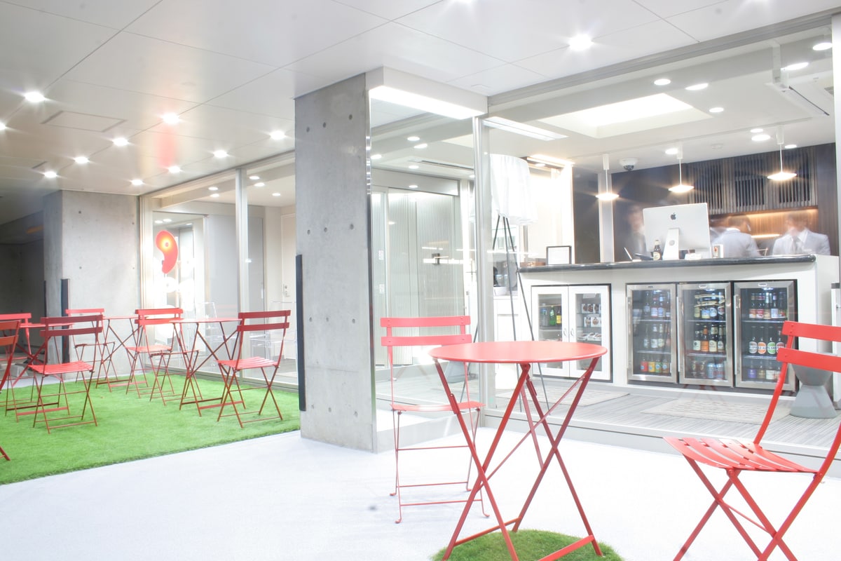 A communal terrace area features red metal tables and chairs set on a green lawn-like surface. On the right, a modern bar is visible with glass doors showcasing various beverages, illuminated by warm overhead lights.