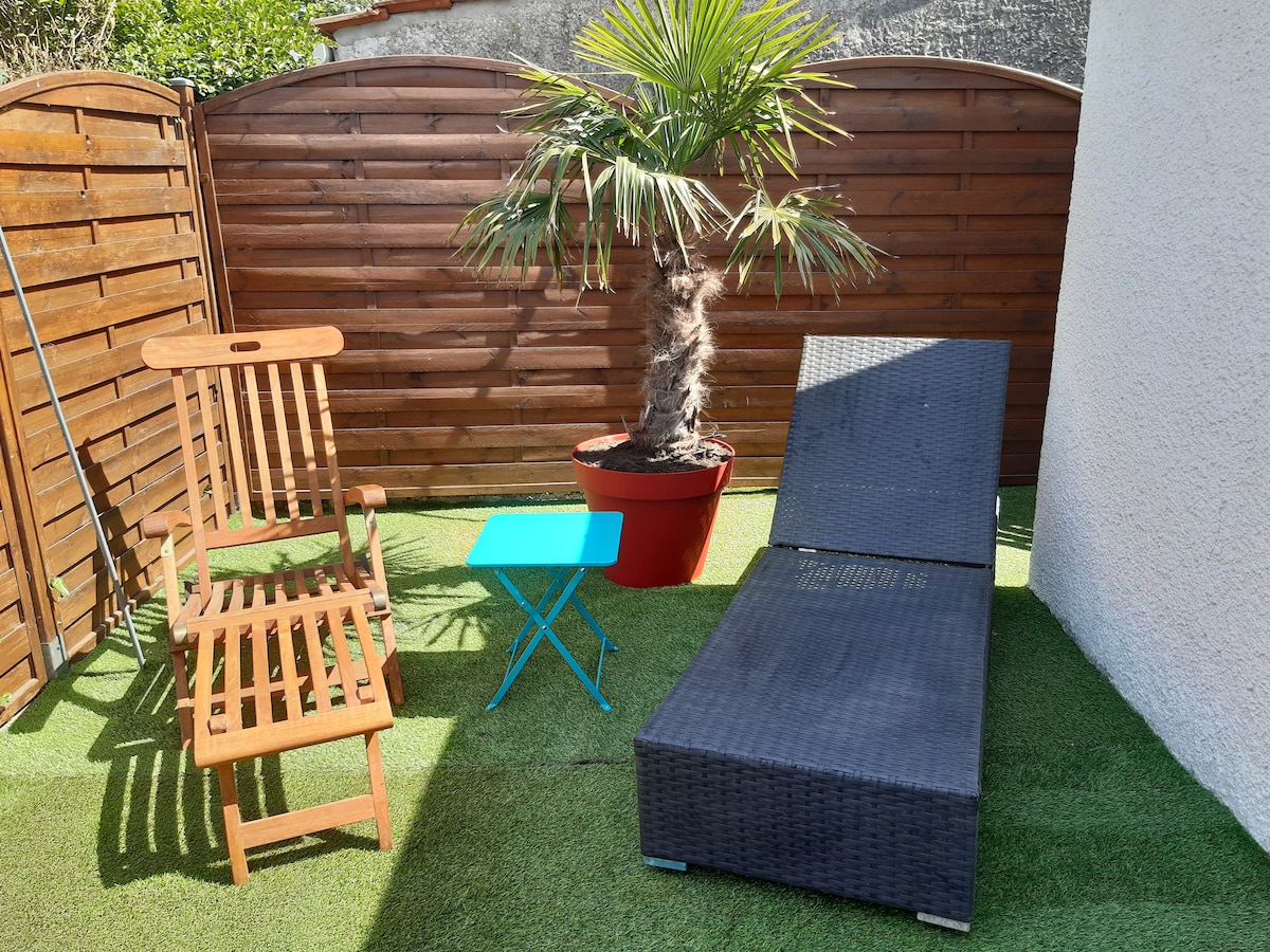 A private courtyard features a tropical palm tree in a vibrant planter. A reclining lounge chair and a wooden chair are positioned on artificial grass, accompanied by a small blue side table. The space is enclosed by wooden fencing, offering privacy for relaxation.