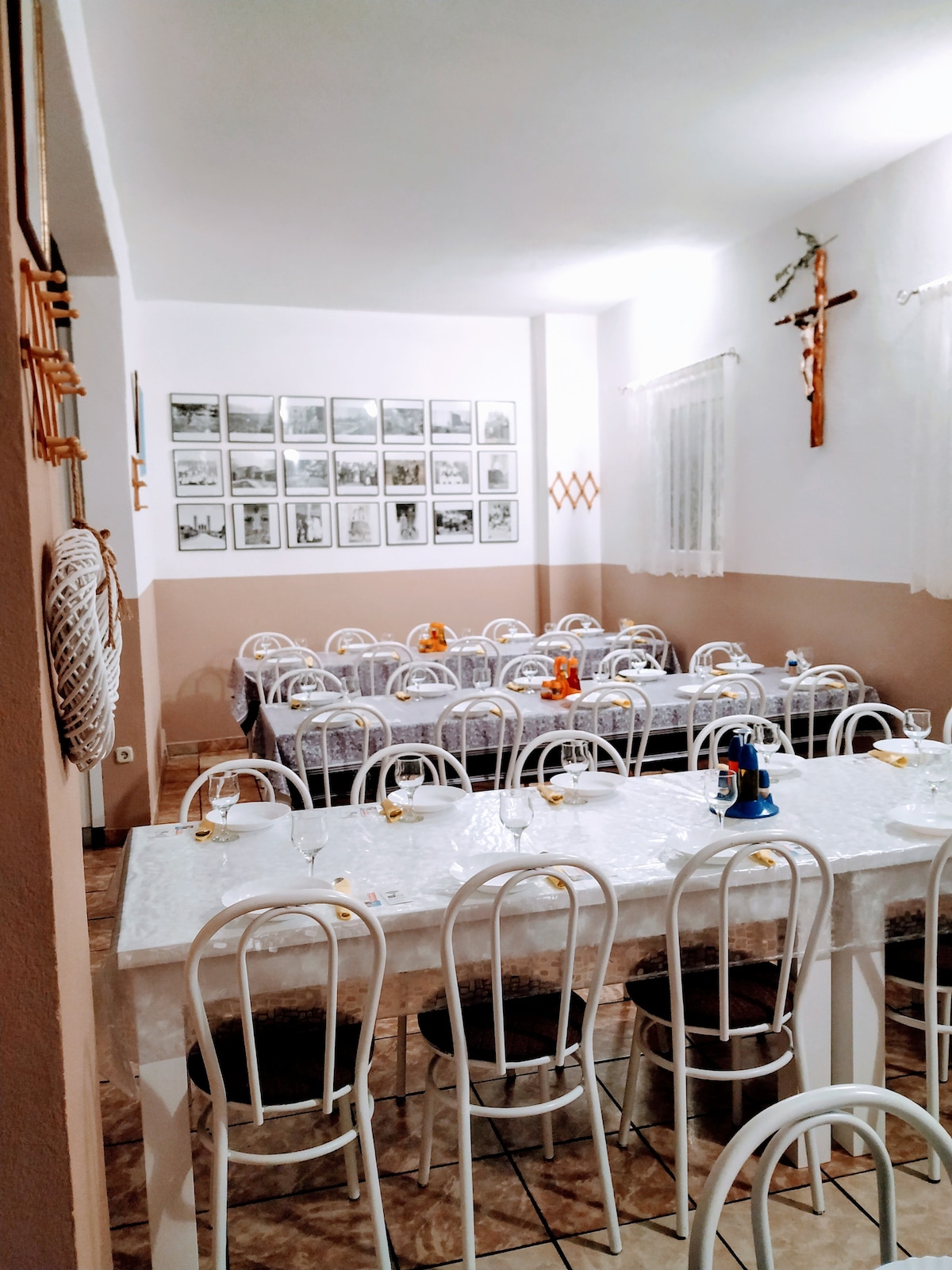 A spacious dining area is arranged with multiple long tables set with white tablecloths and simple place settings. Framed photographs adorn the walls, and a wooden cross is mounted above. Soft lighting creates a calm ambiance, ideal for group dining experiences.