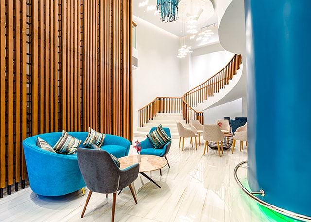 A bright lobby area features stylish blue and gray seating arrangements, complemented by wooden column accents. Soft lighting highlights the contemporary design of the space, while a winding staircase leads to the upper level, creating an inviting atmosphere for guests.