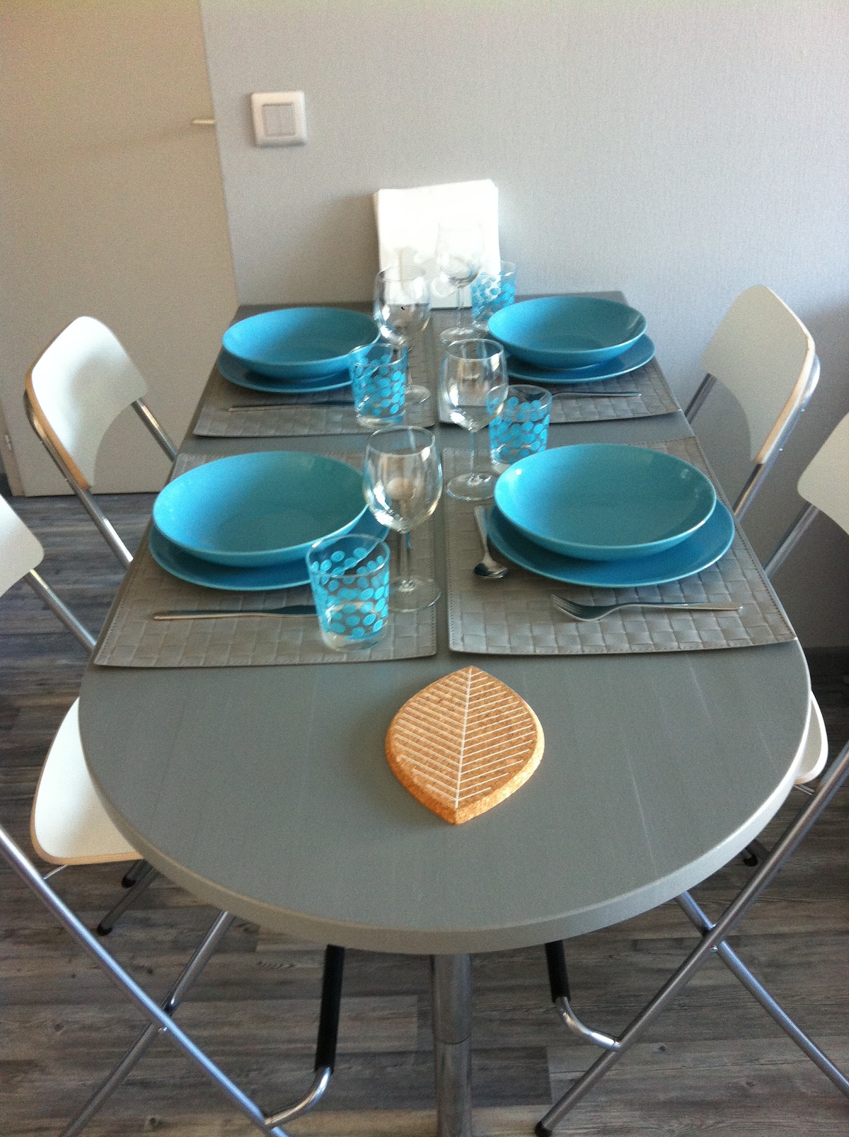 A dining table is set with turquoise dinnerware, including plates and glasses, creating a cohesive look. Silver placemats and a decorative wooden leaf centerpiece enhance the presentation. Six modern chairs are arranged around the table, inviting seating for guests.
