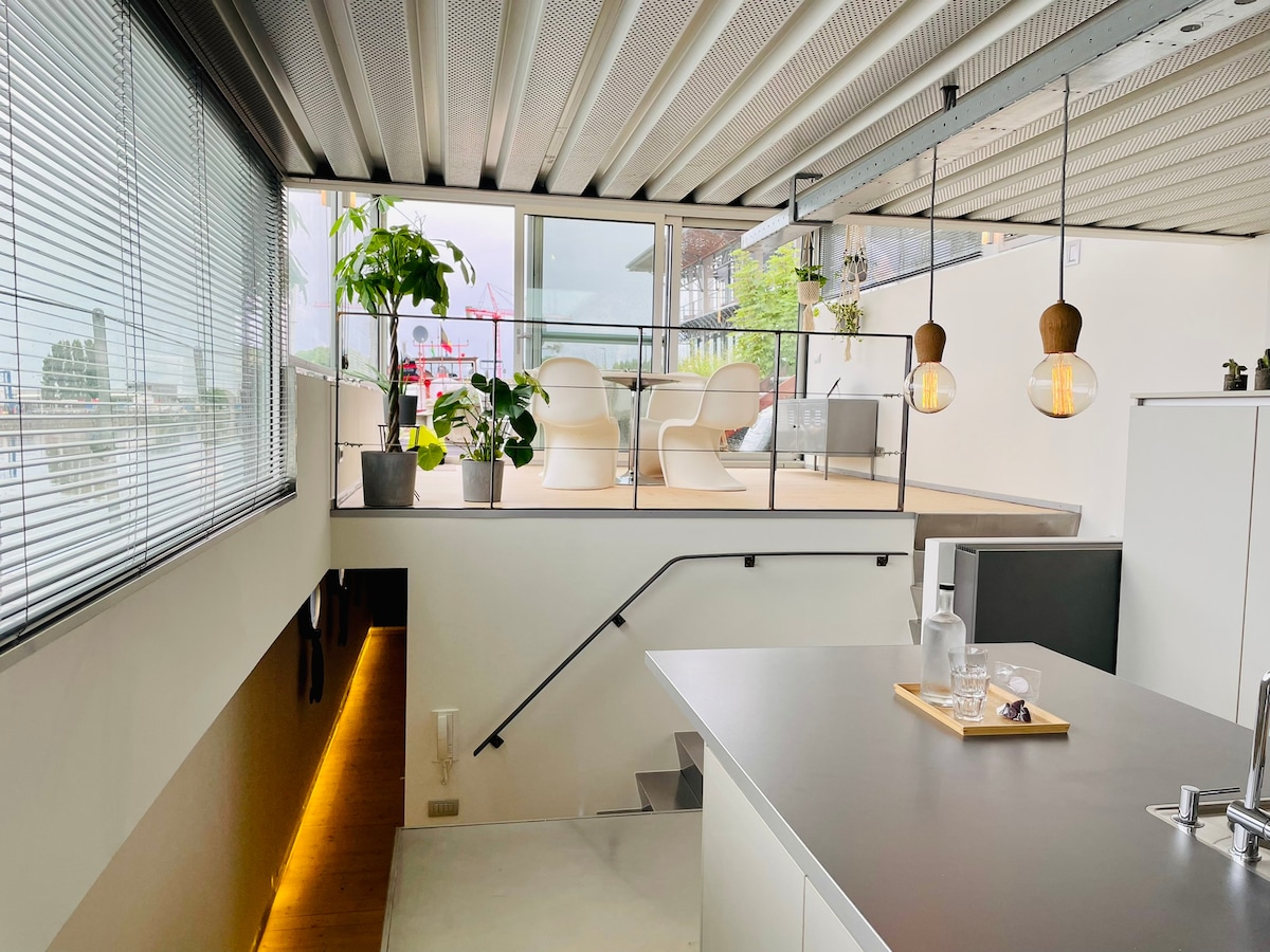An inviting interior space features a sleek kitchen with a central island, illuminated by pendant lights. A staircase leads to a bright area with seating, surrounded by large windows offering views of the outside greenery and waterfront activities in the distance.