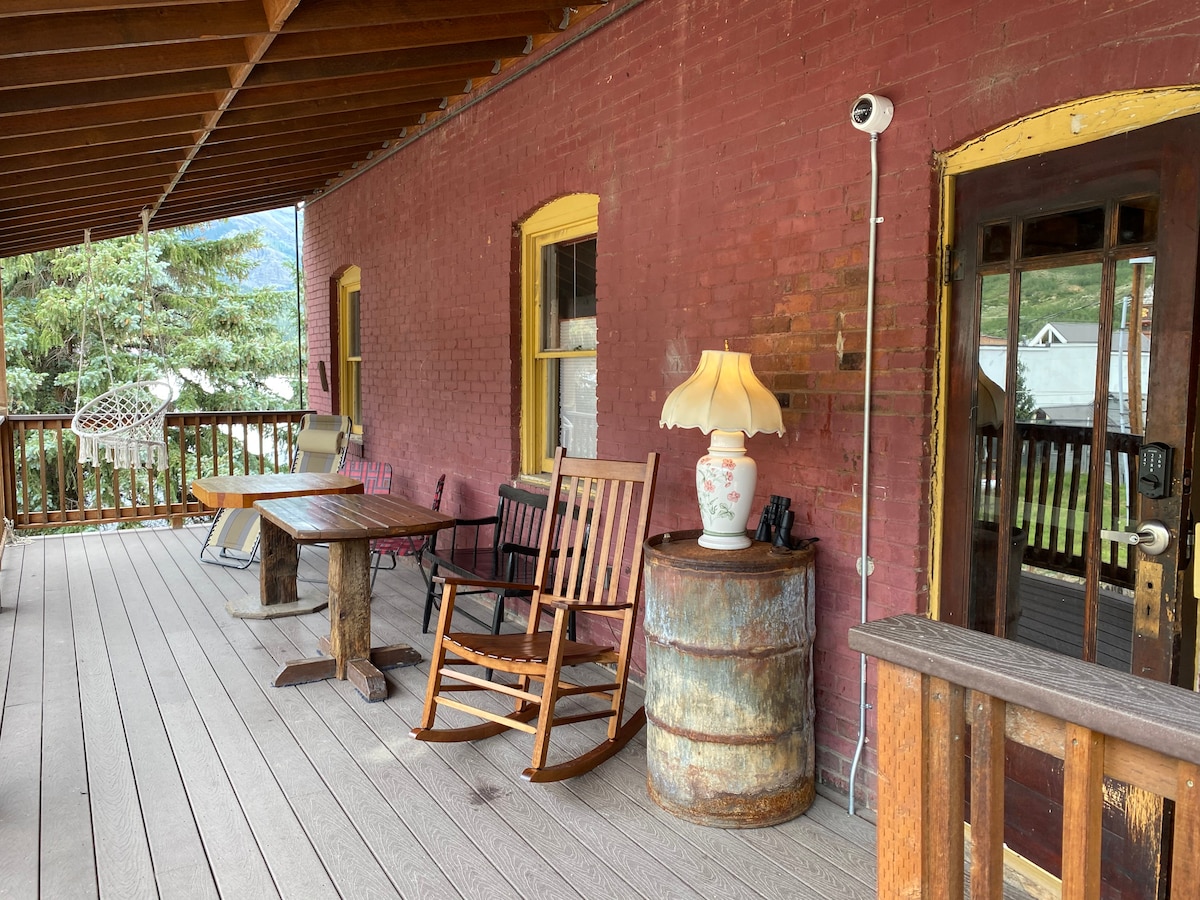 A spacious deck area features wooden flooring and a mix of seating options, including a rocking chair and a table. A decorative lamp sits atop a barrel, contributing to the inviting outdoor space surrounded by trees and a view of the landscape.