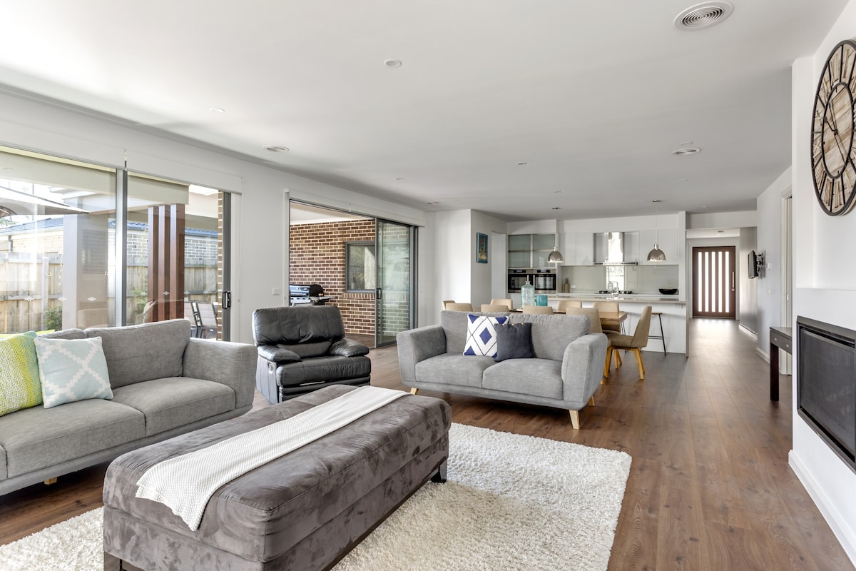 A spacious living area is highlighted by large windows that fill the room with natural light. Modern sofas are arranged around a central ottoman on a soft rug, with a view of the open-plan kitchen and dining area visible in the background.