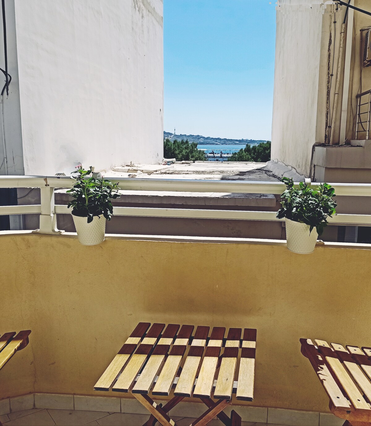 A small balcony is visible, featuring two wooden folding chairs and a table. Potted green plants hang on the railing, with a view of the sea in the background, framed by neighboring buildings. The clear sky enhances the bright, open atmosphere.