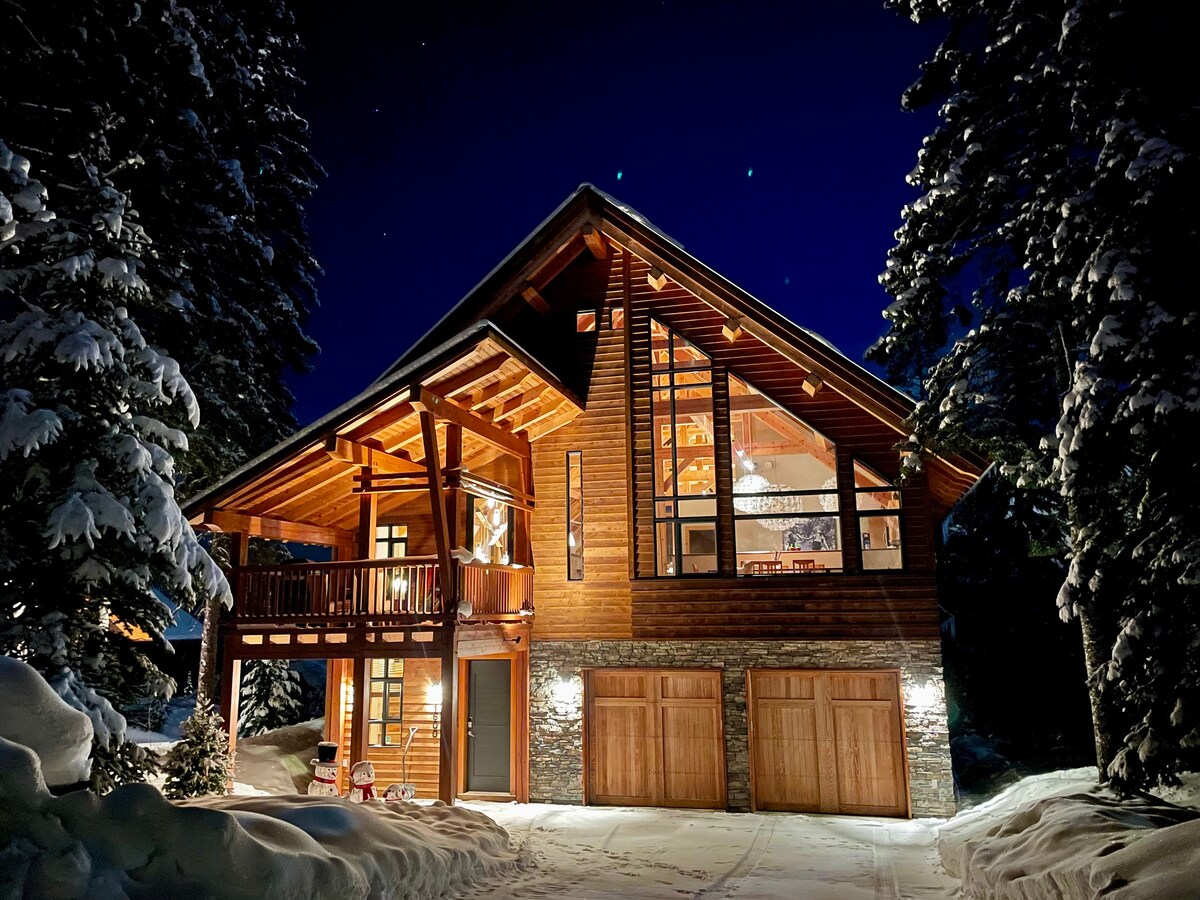 The timber frame chalet stands elegantly under a night sky, highlighted by a warm glow from its windows. Snow-covered trees surround the entrance, while the façade features a combination of wood and stone, contributing to its rustic charm. Two wooden garage doors complete the inviting exterior.