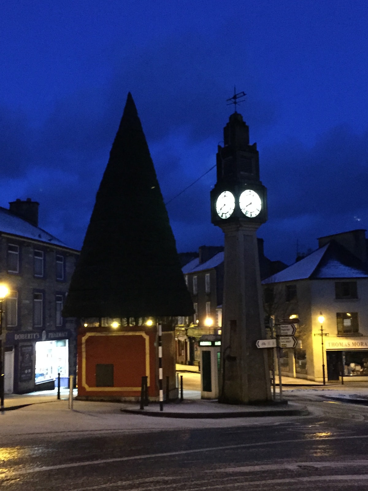 A tall clock tower stands prominently in the center of a town square, illuminated by streetlights. A large conical tree is positioned nearby. The sky exhibits deep blue hues, suggesting twilight. Buildings line the square, displaying their facades and signs.