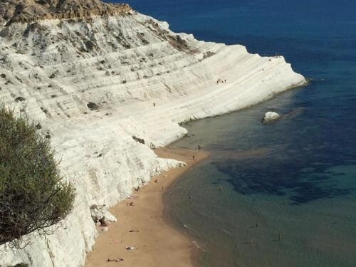 A view of a sandy beach juxtaposed against striking white cliffs, with gentle waves lapping at the shore. Sunbathers dot the beach area, while the clear blue sea stretches into the distance, highlighting the natural beauty of the coastline.