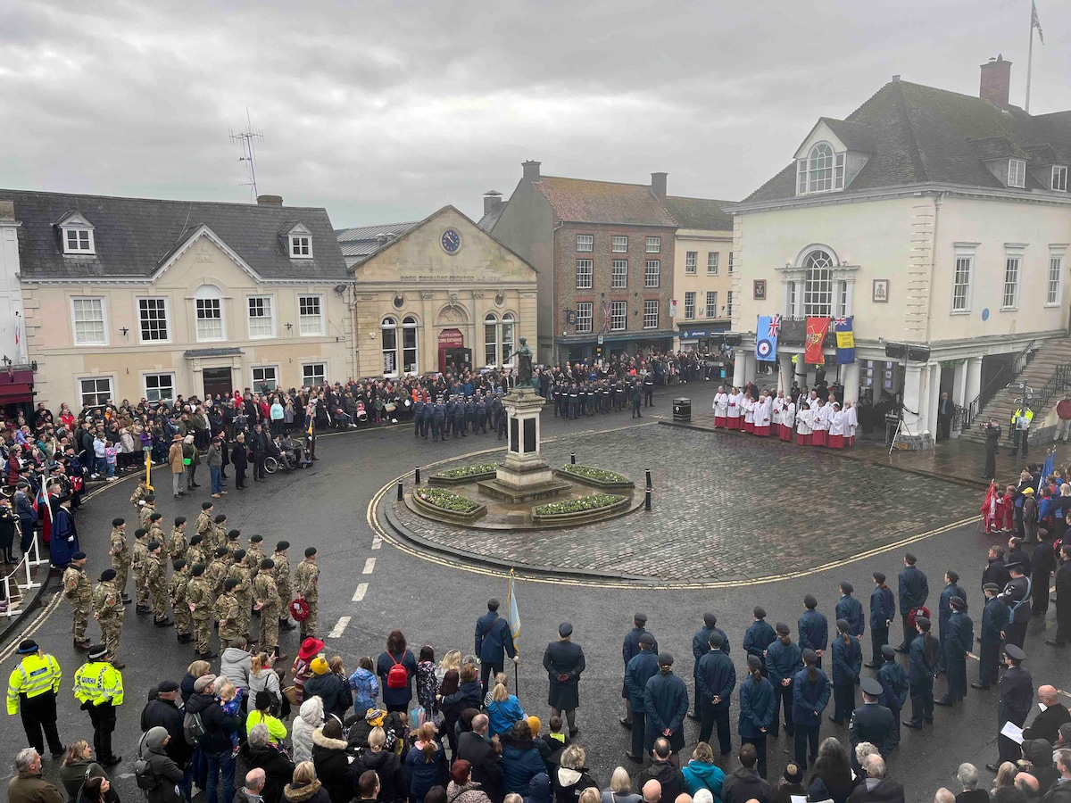 A lively town square is depicted, filled with a diverse crowd gathered for a ceremony. Uniformed groups stand in formation around a central fountain, flanked by historic buildings. Flags and banners add color to the scene, under a gray, overcast sky.
