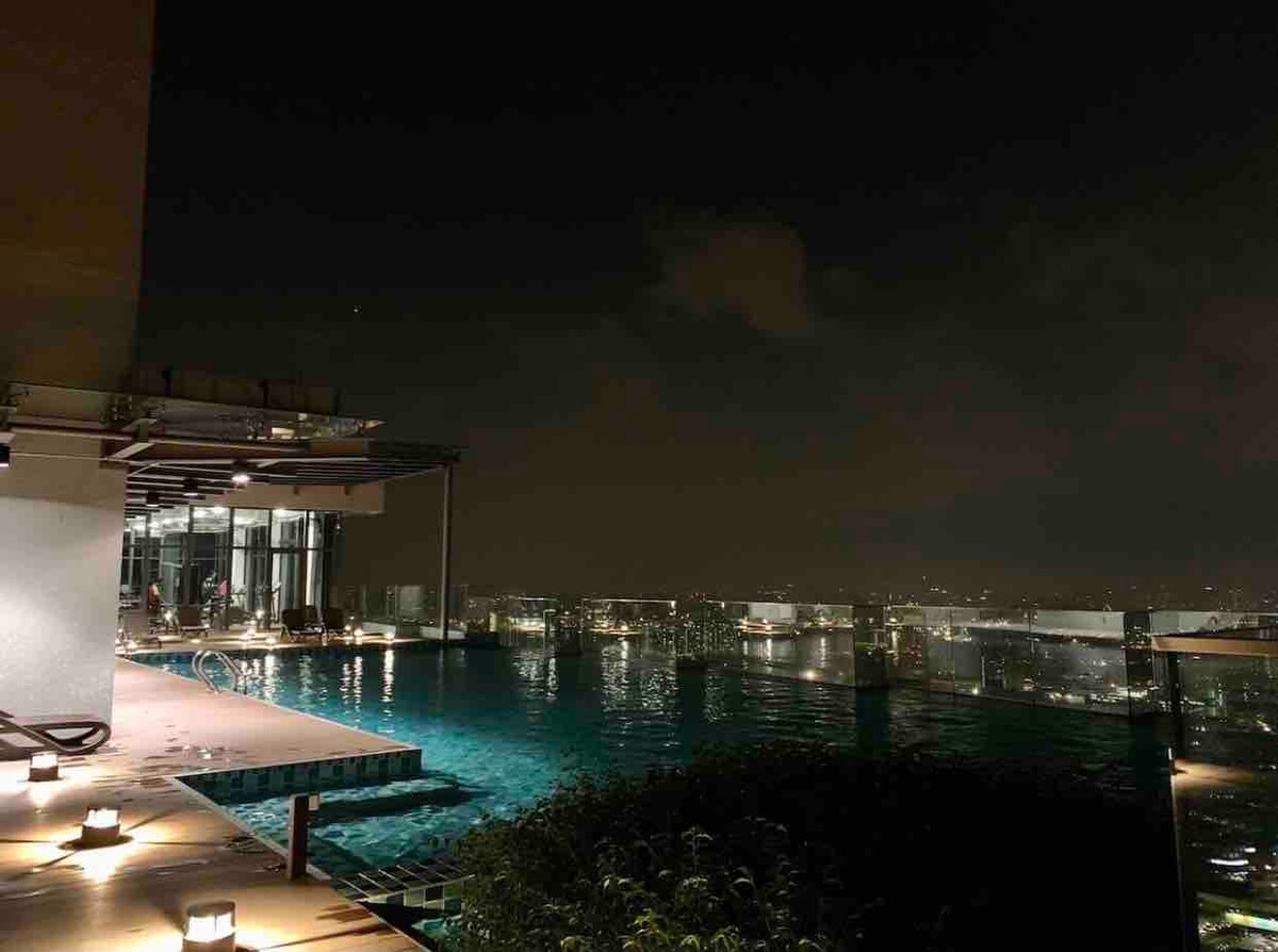 An infinity swimming pool is illuminated at night, offering sweeping views of the city skyline. The pool edge appears to blend into the horizon, and surrounding deck areas are adorned with soft lighting, creating a serene atmosphere for evening relaxation.