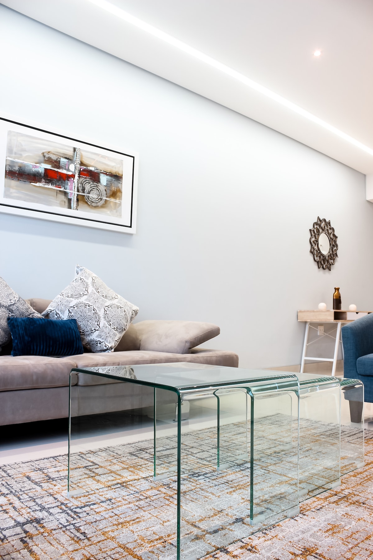 A modern living area features a soft gray sofa with decorative pillows and a reflective mirror on the wall. A glass coffee table with a sleek design stands on a patterned rug, complemented by soft lighting from above.