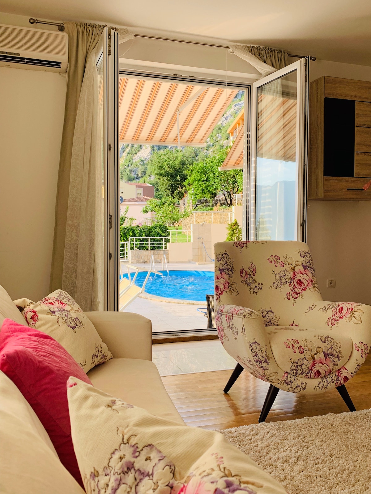 A cozy living area features a floral-patterned armchair facing large glass doors that open to reveal a poolside view. Natural light floods the space, which is adorned with soft, neutral furnishings and decorative pillows, creating a comfortable ambiance.
