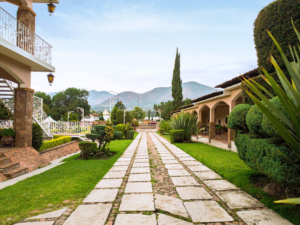 A winding stone path leads through lush green lawns and manicured gardens, bordered by neatly trimmed shrubs and trees. In the background, hills are visible under a clear sky, contributing to a serene atmosphere. The pathway connects various outdoor spaces, promoting relaxation.