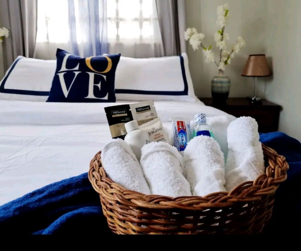 A neatly arranged basket filled with rolled white towels and essential toiletries is positioned on the edge of a spacious bed. The bed features crisp white linens and a decorative pillow with the word 'LOVE.' Soft lighting from a nearby lamp enhances the calm ambiance.