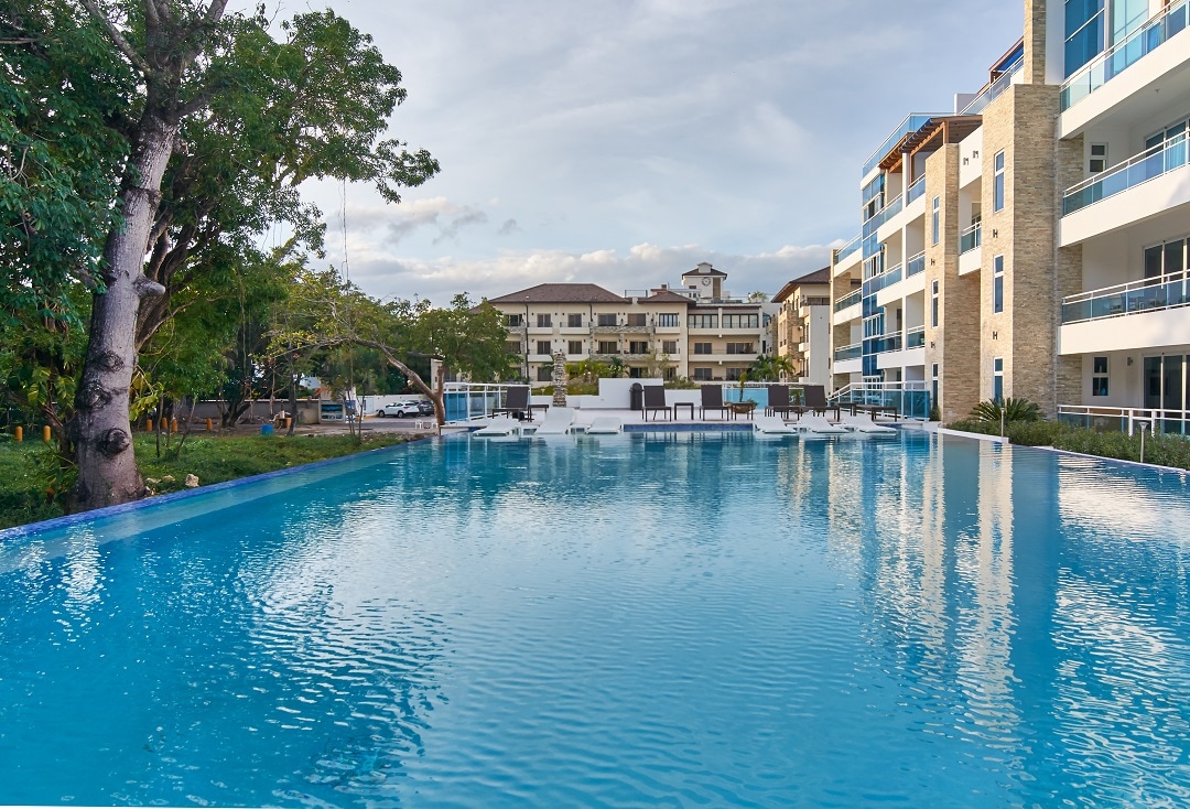A tranquil pool area is showcased, featuring clear blue water that reflects the surrounding greenery. Sun loungers are positioned along the edge, complemented by a vibrant landscape of trees. The modern condominium building stands nearby, providing a contemporary setting.