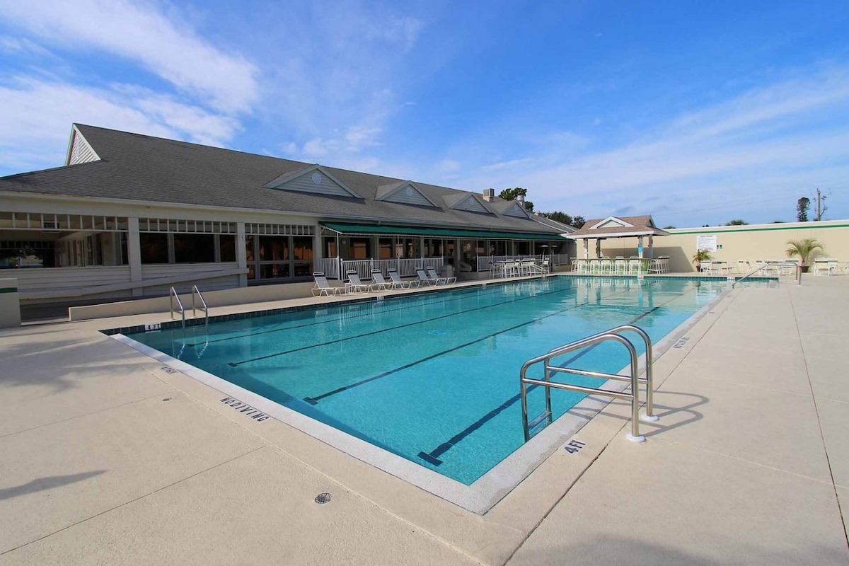An expansive outdoor pool is featured, surrounded by a spacious deck with lounge chairs. The pool's clear blue water reflects the sky, while a multi-panelled building with large windows is visible in the background. The atmosphere is tranquil and inviting.