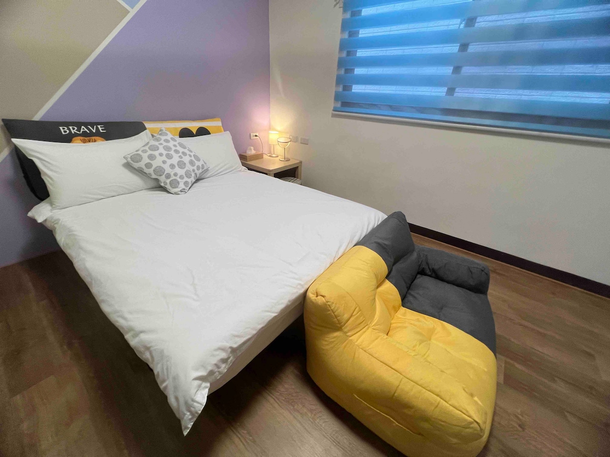A neatly arranged sleeping area features a spacious bed with white linens and a decorative pillow. A soft, dual-toned floor chair in gray and yellow is positioned beside the bed. Natural light filters through a modern window with sheer shades.
