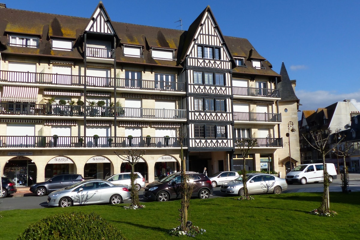 A charming residence features a traditional Norman architectural style, displaying wooden beams and balconies. The building is situated on a bright plaza with manicured grass and trees, alongside parked cars. The surrounding area showcases shops and cafes, contributing to a welcoming environment.