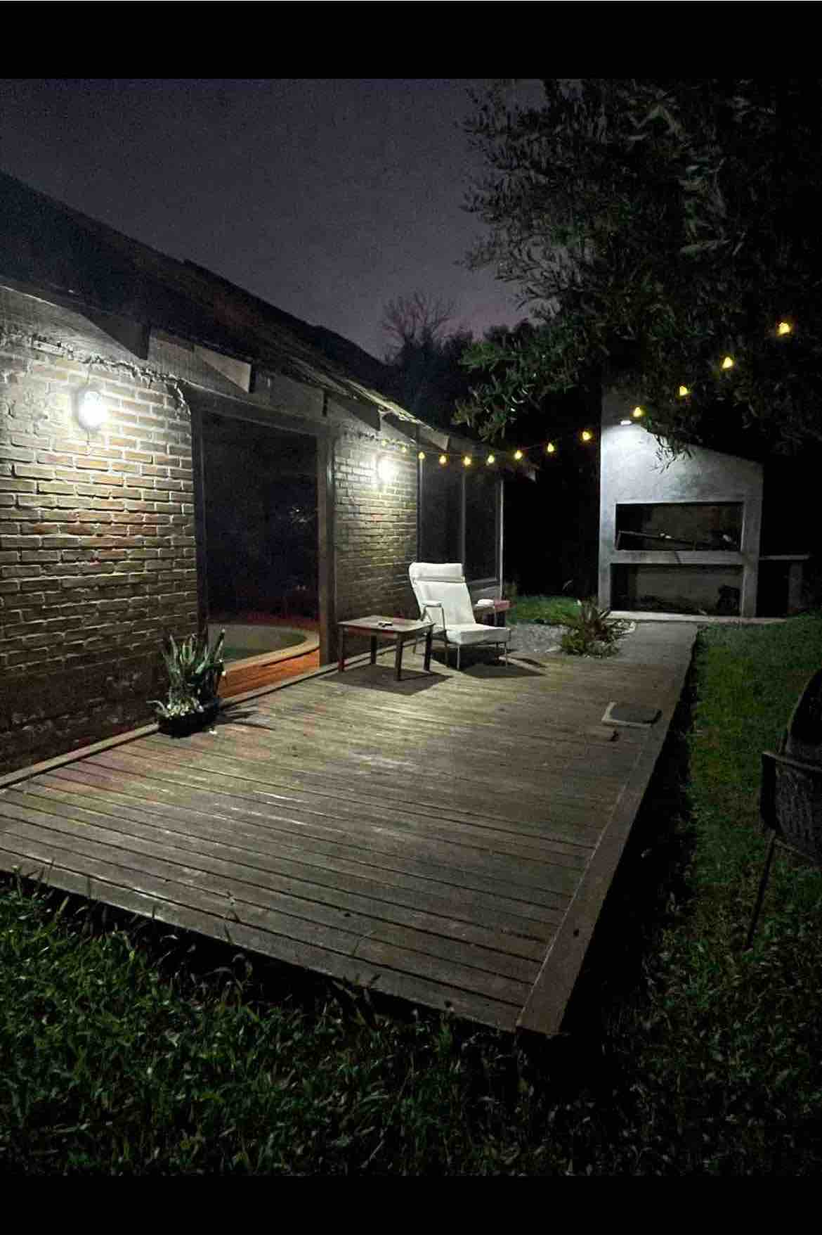 A wooden deck is highlighted by soft outdoor lighting, showcasing a seating area with a chair and a small table. A barbecue grill stands nearby, surrounded by lush greenery. The area exudes a peaceful ambiance, ideal for relaxation in a natural setting.