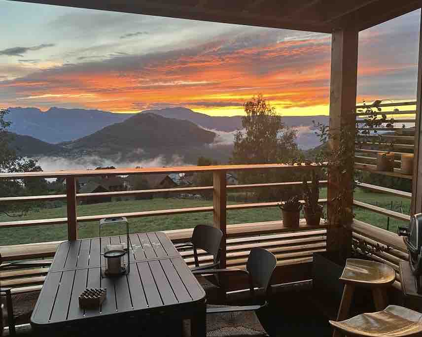 A covered terrace features a wooden dining table surrounded by chairs, offering a panoramic view of the valley and mountains at sunset. The sky displays hues of orange and pink, with clouds chilling in the valleys below. Potted plants add natural elements to the setting.