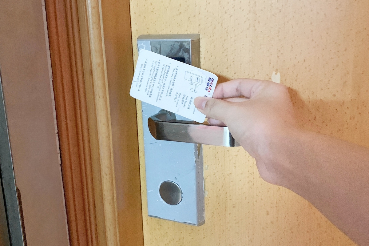 A hand is shown inserting a key card into a modern door lock. The door features a natural wood finish with a metallic handle and a circular keyhole. Clear signage is visible on the card, which is used for accessing the room.