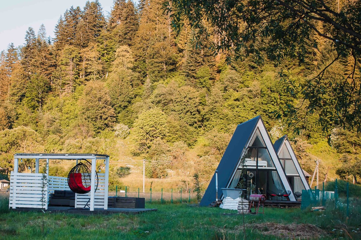 A unique A-frame structure is situated amidst lush green hills, surrounded by trees. A cozy outdoor seating area, featuring a circular hanging chair, complements the architectural design. The serene landscape invites a peaceful retreat, with open grassy space connecting the living areas to nature.
