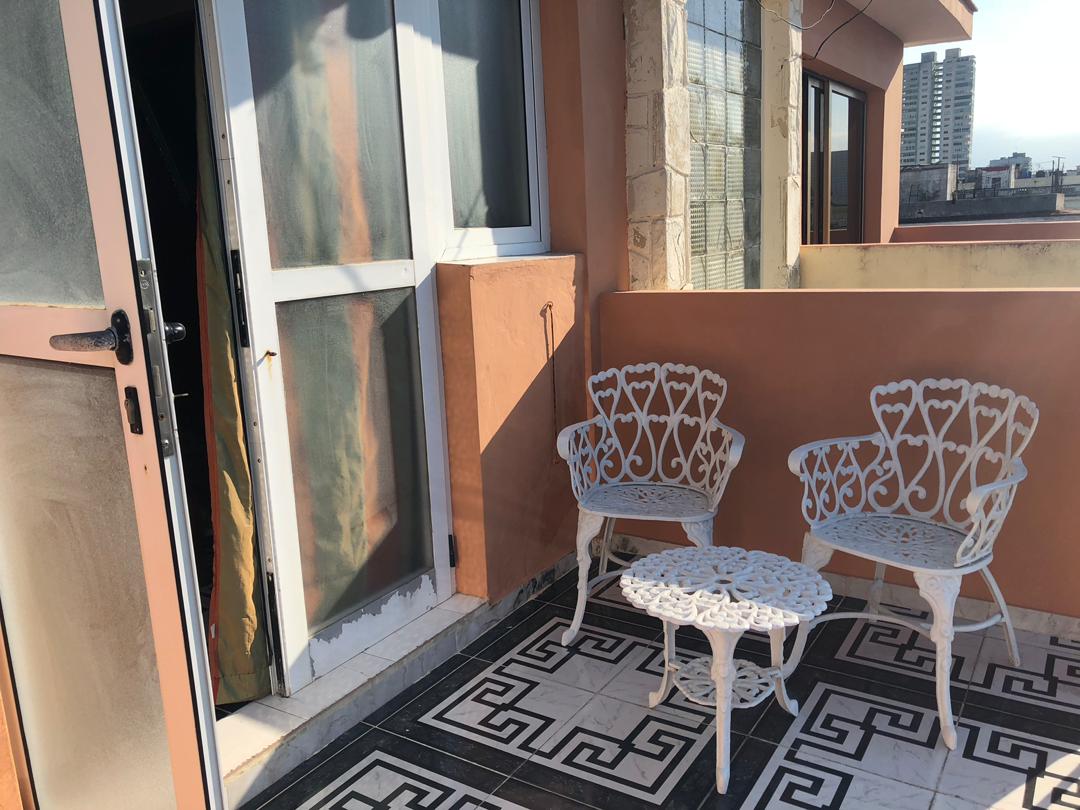 A small terrace features two intricately designed white metal chairs and a matching table, arranged on a patterned tiled floor. Sunlight filters through a nearby door, enhancing the inviting outdoor space with views of the surrounding neighborhood.