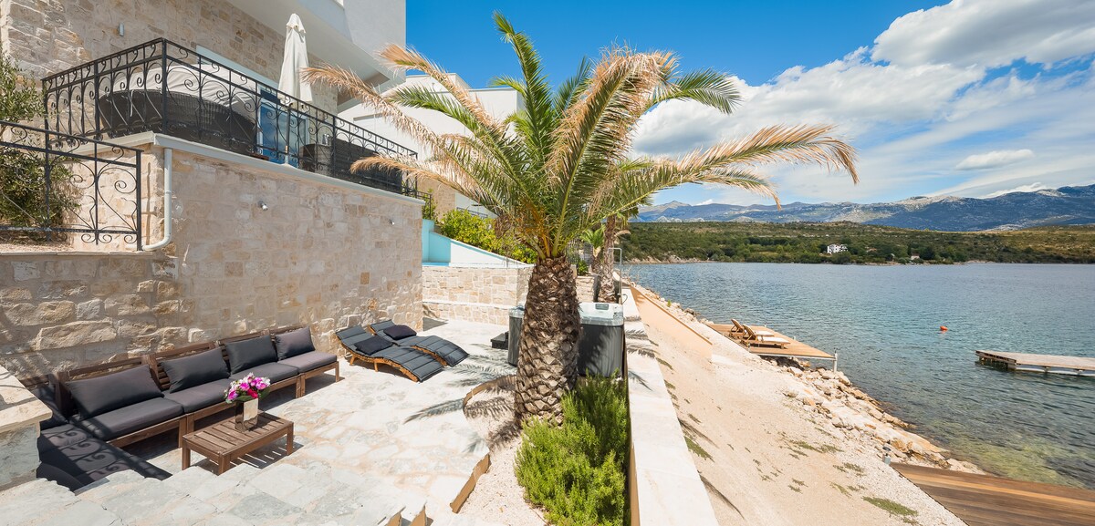 A serene outdoor area borders the water, featuring a palm tree alongside a stone terrace. Comfortable lounge chairs are arranged for relaxation, with a low wooden table holding vibrant flowers. The adjacent beach extends towards calm waters, framed by a backdrop of distant hills and clear skies.
