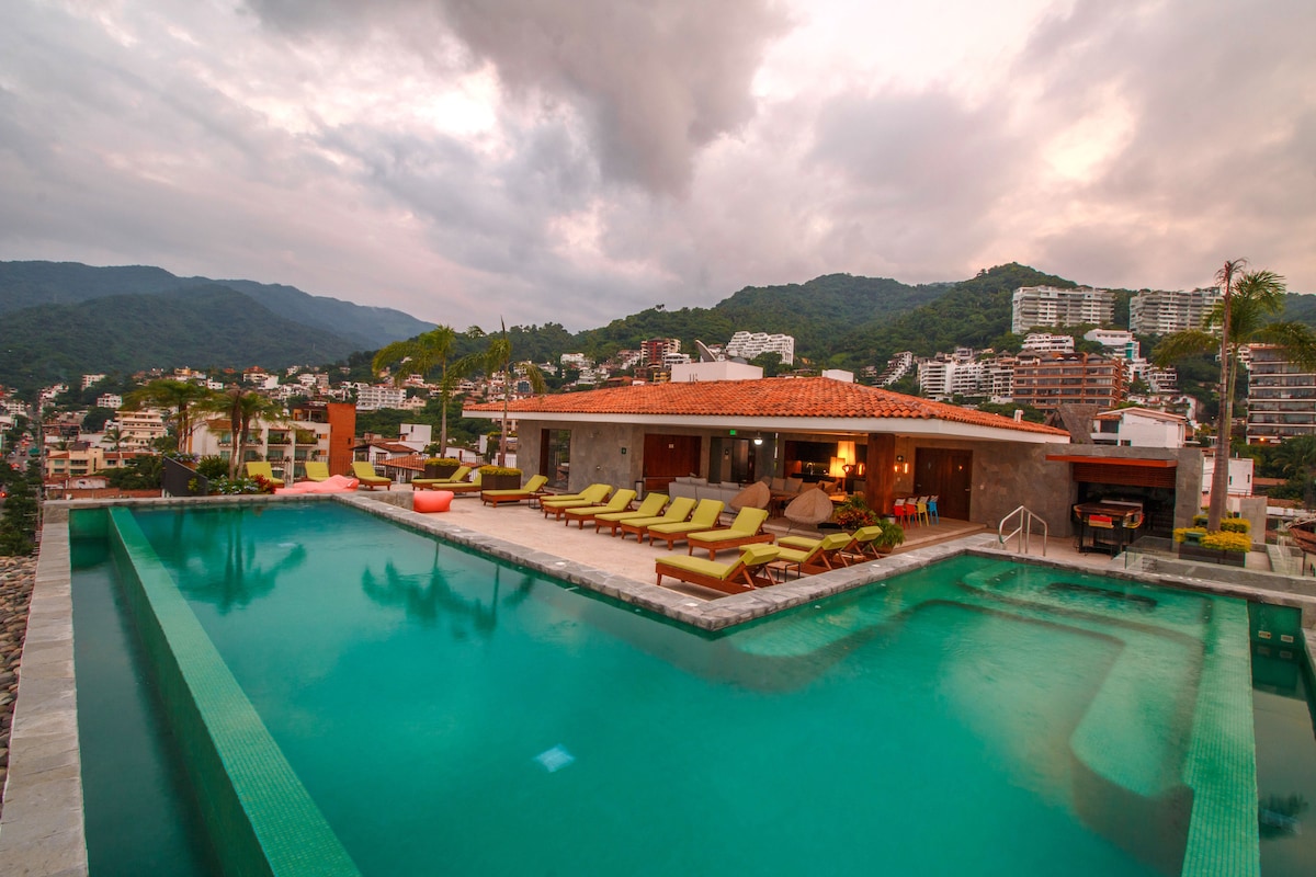 An expansive rooftop pool features an infinity edge on two sides, surrounded by lounge chairs and vibrant umbrellas. Mountains and city buildings provide a scenic backdrop under a cloudy sky, creating a tranquil atmosphere for relaxation.