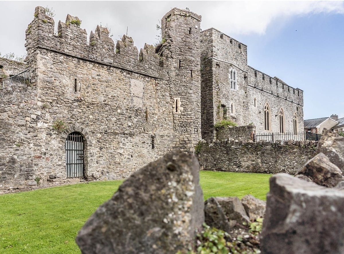 A historic stone castle is depicted, featuring a robust exterior with a mixture of high walls and turrets. The structure is partially surrounded by manicured grass and rocky landscaping, highlighting the castle's enduring presence in the landscape.