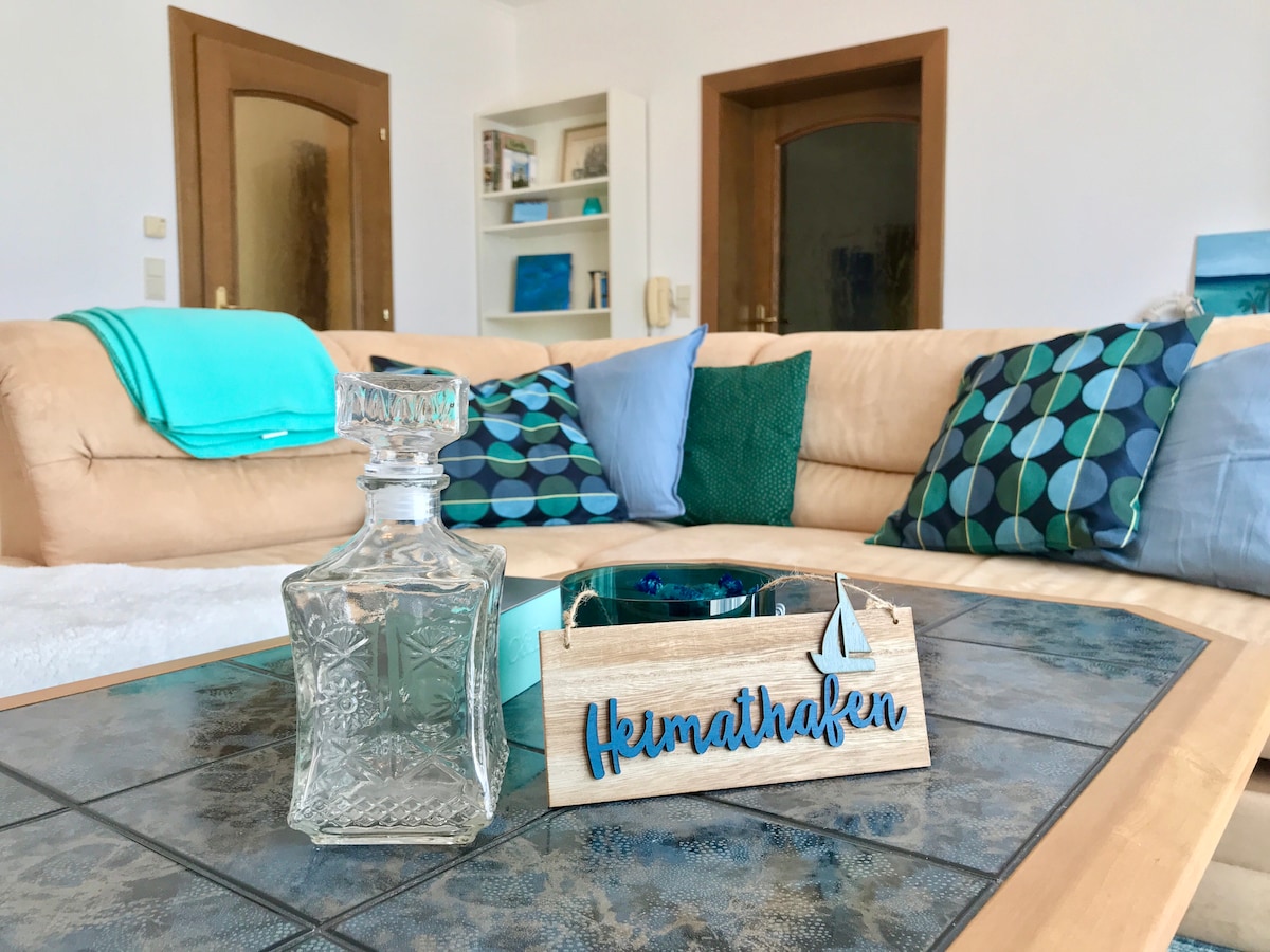 A light-colored leather sofa is adorned with cushions in various shades of blue and green. A glass decanter and a wooden decorative box labeled 'Heimathafen' are placed on a coffee table, enhancing the living area’s welcoming ambiance.