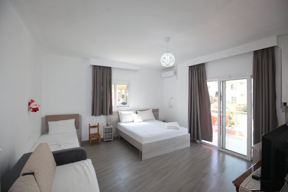 A bright and airy room featuring a spacious bed, along with a single bed and a pull-out sofa. Large glass doors provide access to a terrace, with natural light streaming in through the windows adorned with light curtains.