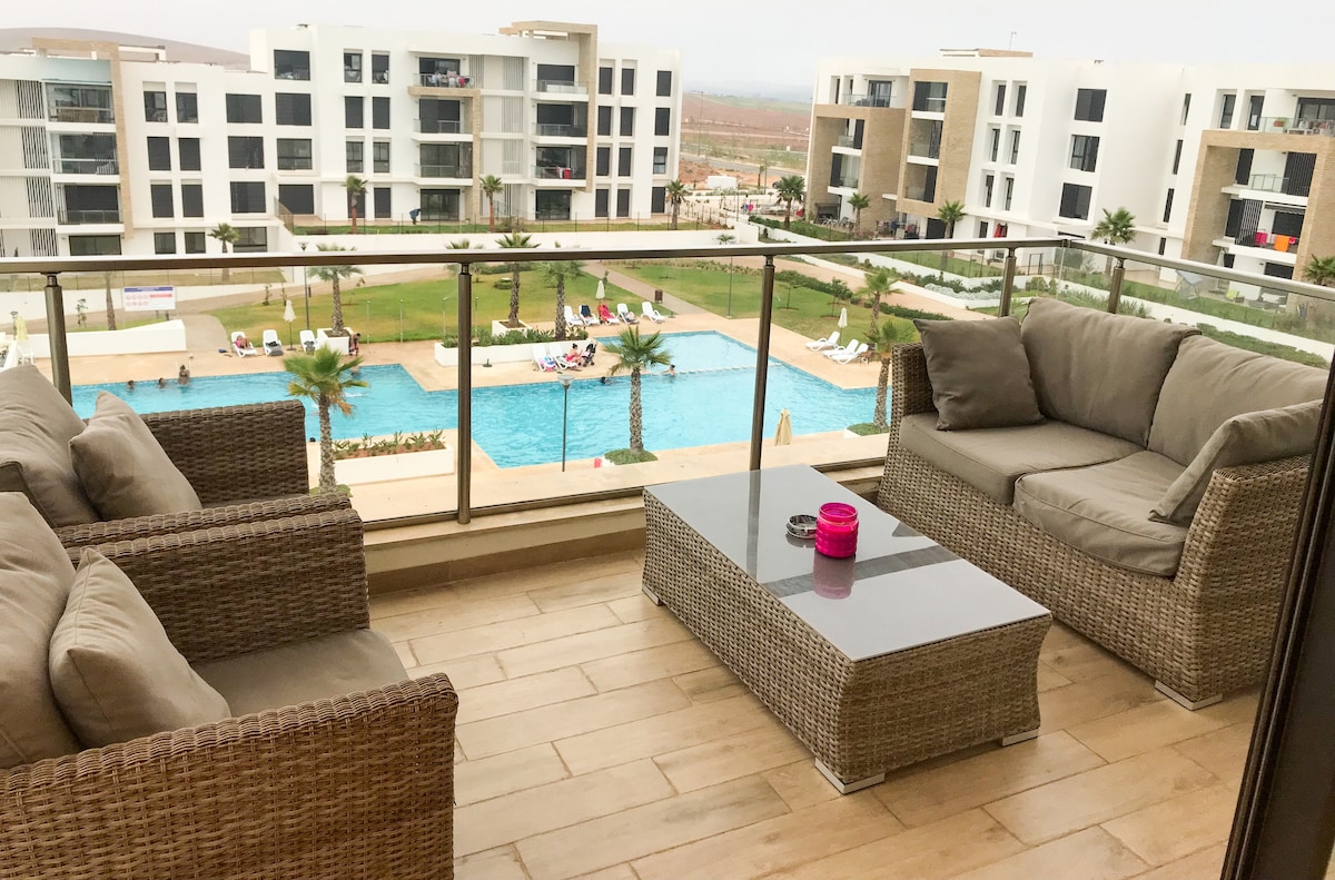 A balcony is furnished with comfortable seating arranged around a coffee table. The view features a large swimming pool surrounded by palm trees and lounge chairs, set against a backdrop of modern residential buildings.