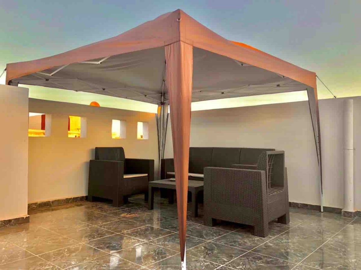 A shaded seating area is provided under a canopy, featuring several comfortable outdoor sofas and a central table. The flooring is covered with polished tiles, and light is filtered through small windows in the surrounding wall, creating a relaxing atmosphere.