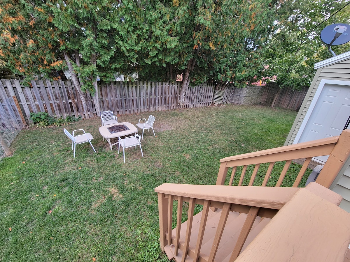 A fenced-in backyard features a well-maintained lawn surrounding a fire pit area. Four white chairs are arranged around a small table, inviting gatherings. Lush green trees provide shade, enhancing the peaceful ambiance.