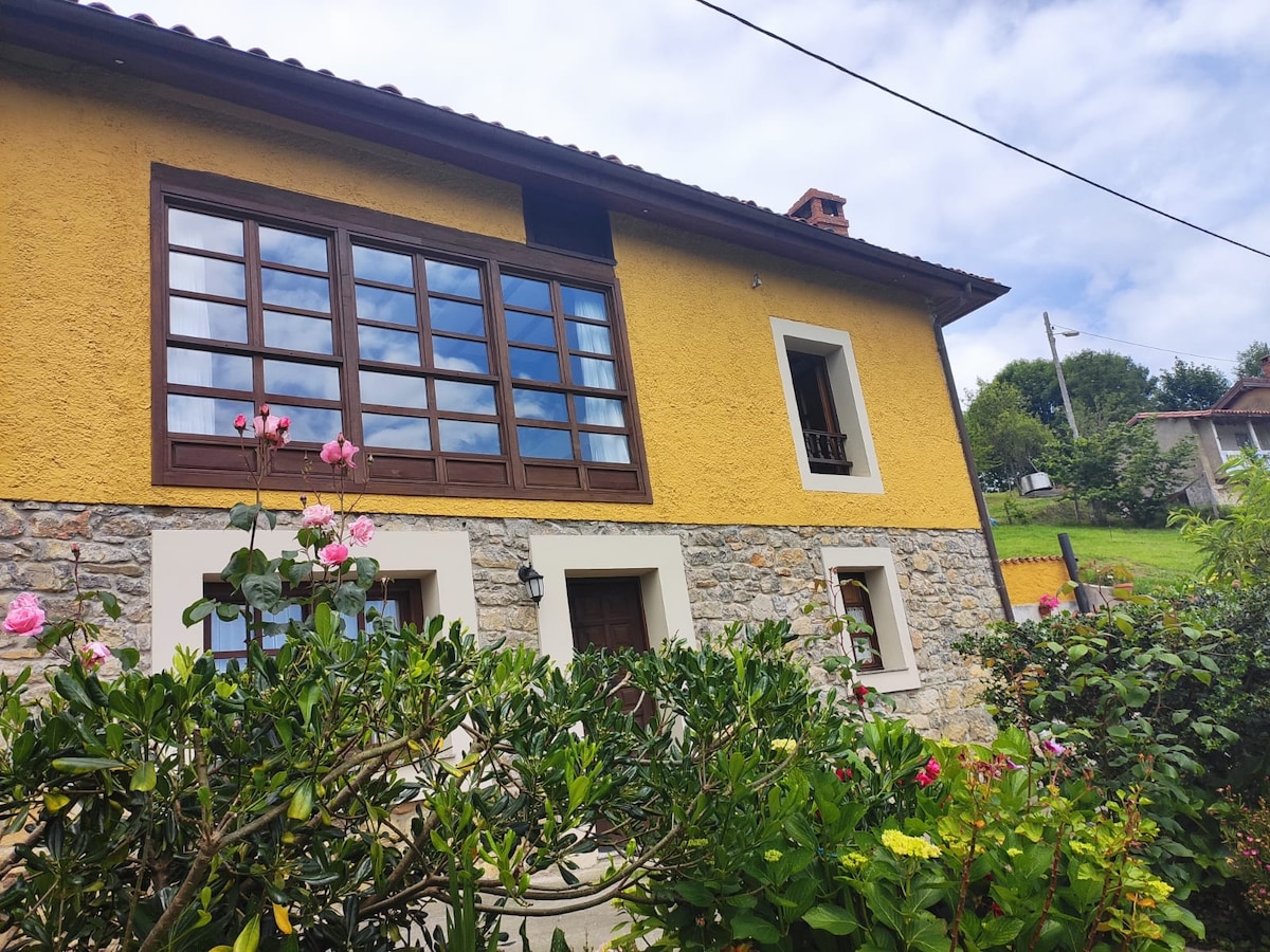 The exterior of a charming house is shown, featuring a yellow wall and stone accents. Large windows with brown frames provide a view of the greenery outside. Colorful flowers in the foreground add a touch of nature to the inviting setting.