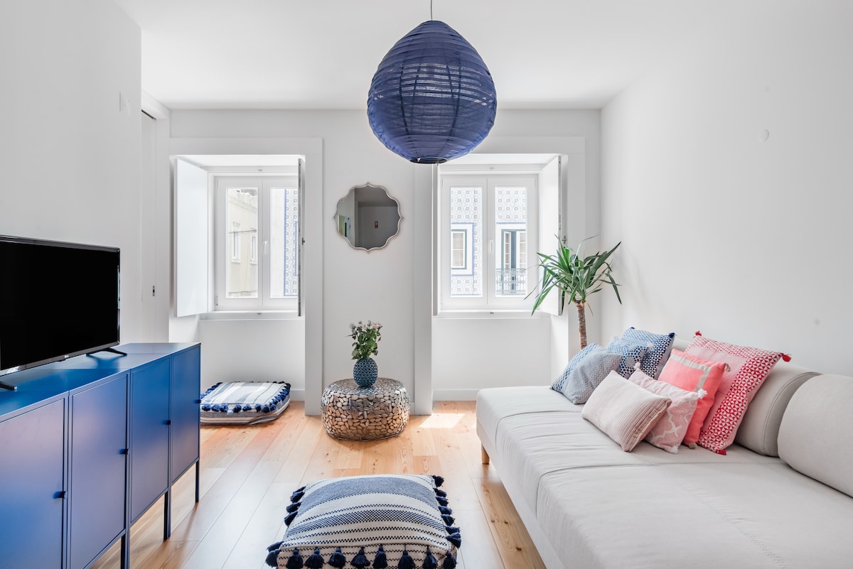 A bright living room features large windows allowing natural light to fill the space. A light-colored sofa is accompanied by colorful cushions. A blue pendant lamp hangs above. A small plant and decorative table add touches of greenery, while a TV stands on a blue cabinet.