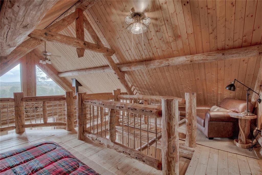 A cozy loft area features wooden beams and a warm wood-paneled ceiling. A spacious bed with a plaid coverlet sits near the railing, overlooking the expanse below. A comfortable seating area is highlighted by a leather chair and a side table, creating a welcoming retreat.